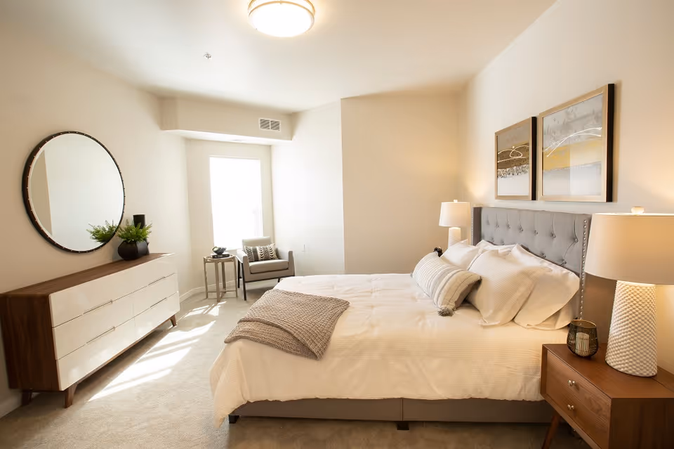 A bright and cozy bedroom with a large bed featuring a tufted gray headboard, white bedding, and multiple pillows. On the right side of the bed is a wooden nightstand with a white textured lamp and a small decorative item. Above the bed are two framed abstract artworks. On the left side of the room is a modern dresser with a round mirror above it and a small plant on top. Near the window at the back is a gray armchair with a patterned pillow and a small side table with a decorative bowl. The room has light-colored walls and carpeted flooring.