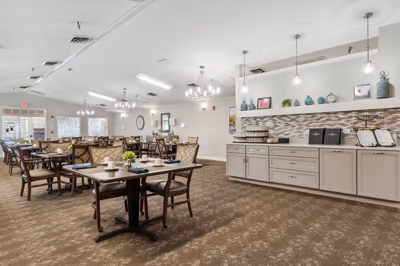 A spacious dining room with multiple tables and chairs arranged neatly. Each table is set with white cups, saucers, and blue napkins. The room features beige walls, carpeted flooring, and modern light fixtures hanging from the ceiling. A long counter with cabinets and decorative items is visible on the right side of the image.