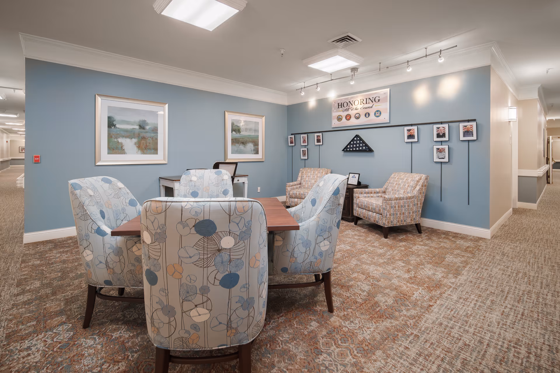 A cozy seating area in a senior living facility with a wooden table surrounded by four upholstered chairs featuring a floral pattern. Two additional patterned armchairs are positioned against a blue wall adorned with framed pictures and a display honoring veterans, including a folded American flag in a triangular case. The space is well-lit with ceiling lights and has a carpeted floor with a patterned design.