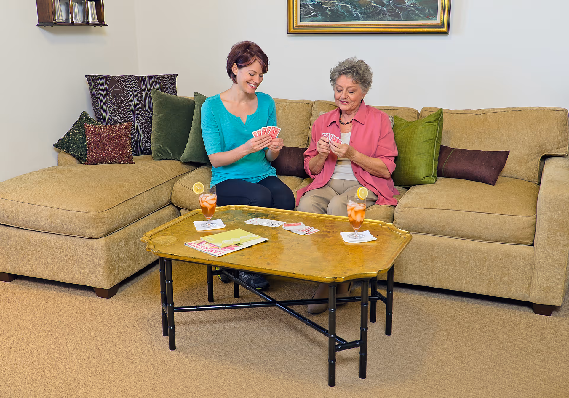 Two women sitting on a beige sectional sofa in a living room, playing cards and smiling. A wooden coffee table in front of them holds playing cards, two glasses of iced tea with lemon slices, and some papers. The room has decorative pillows on the sofa and a framed painting on the wall behind them.
