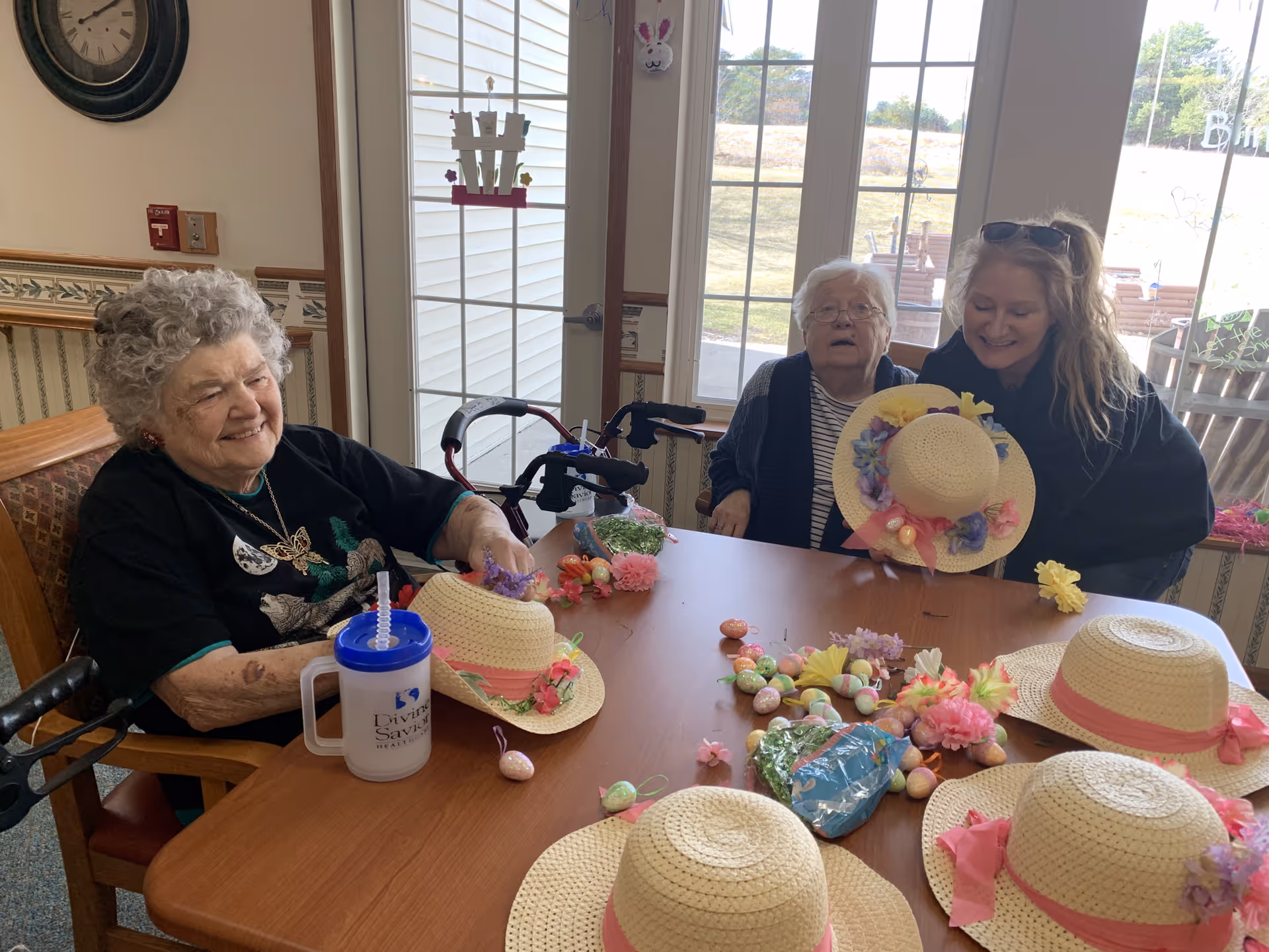 Two elderly women and a younger woman sitting around a table in a senior living facility, decorating straw hats with flowers and Easter-themed decorations. The room has large windows with a view of the outdoors and a clock on the wall.