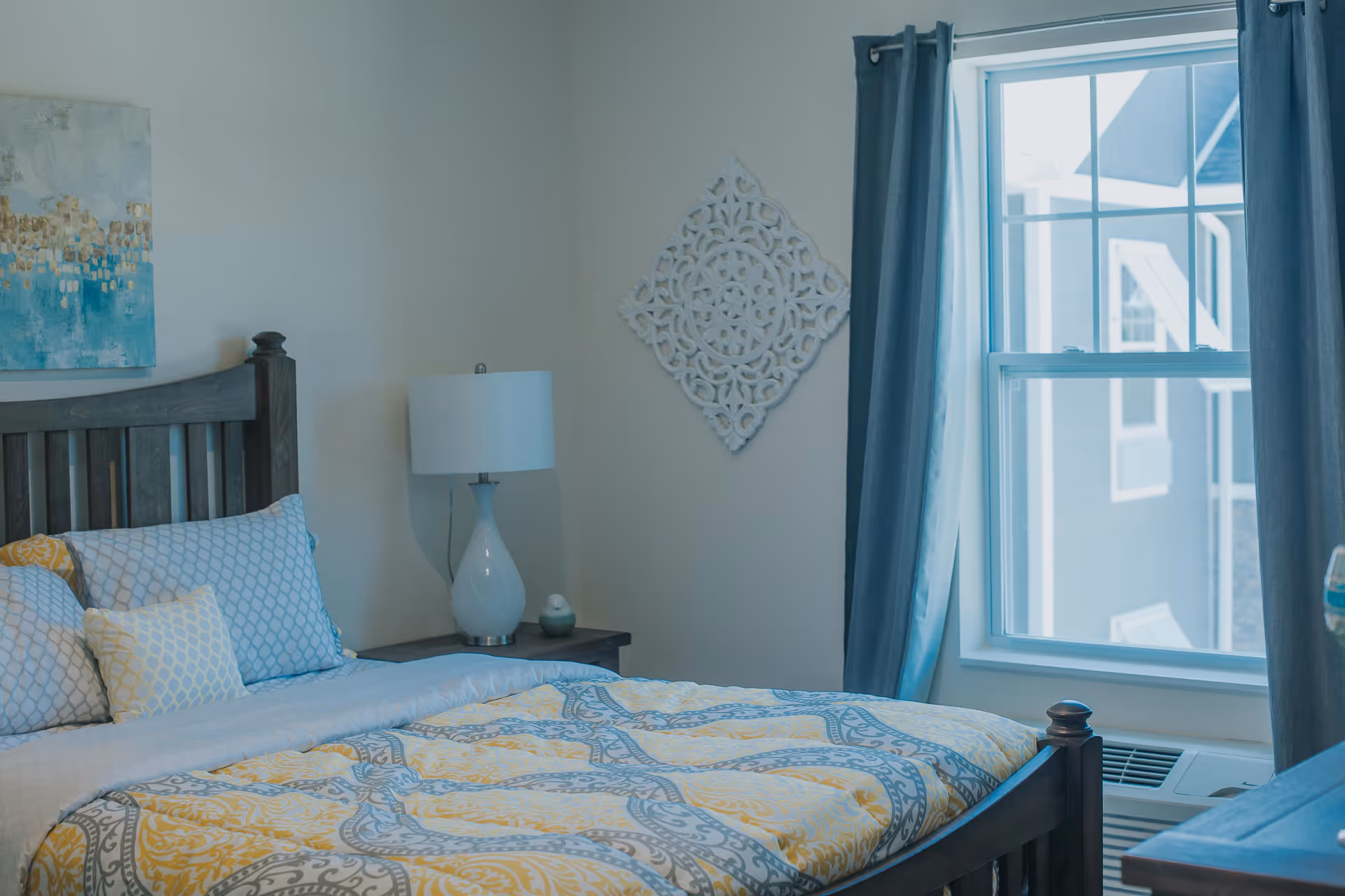 A bedroom with a wooden bed frame, a bed made with patterned bedding in yellow, gray, and white colors, a bedside table with a white lamp, a decorative wall piece, and a window with blue curtains letting in natural light.