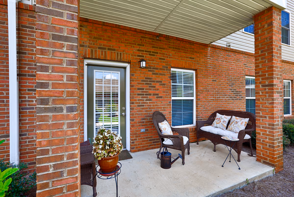 Covered outdoor patio area with brick walls, a glass door with white blinds, two windows with white blinds, a brown wicker loveseat and chair with floral cushions, a small round table, a potted plant on a stand, and a watering can on the concrete floor.