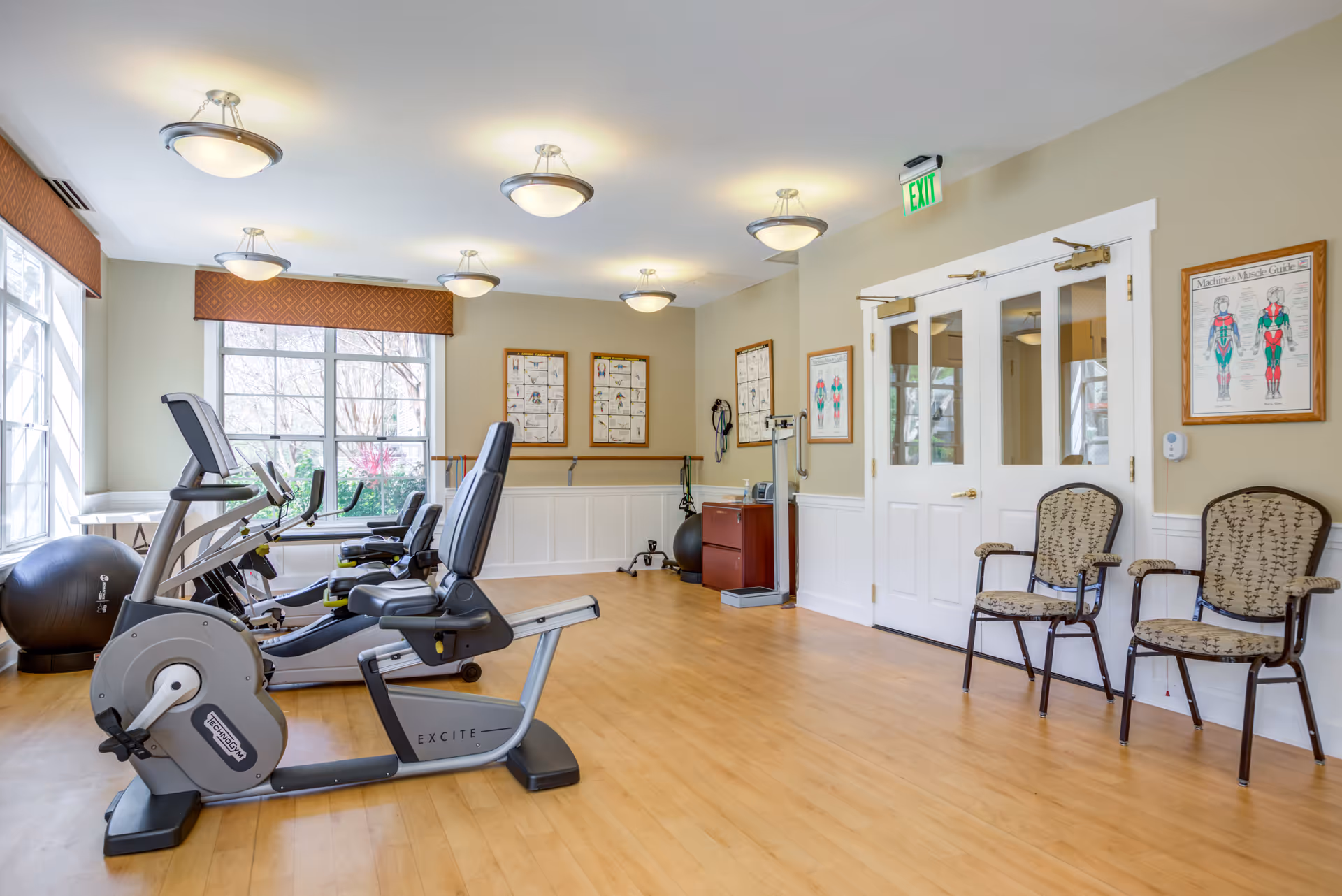 A bright exercise room with large windows letting in natural light. The room contains two stationary exercise bikes, an exercise ball, and various fitness equipment. There are framed anatomical charts on the walls and two patterned chairs near a set of double doors. The floor is light wood and the walls are painted a soft beige.
