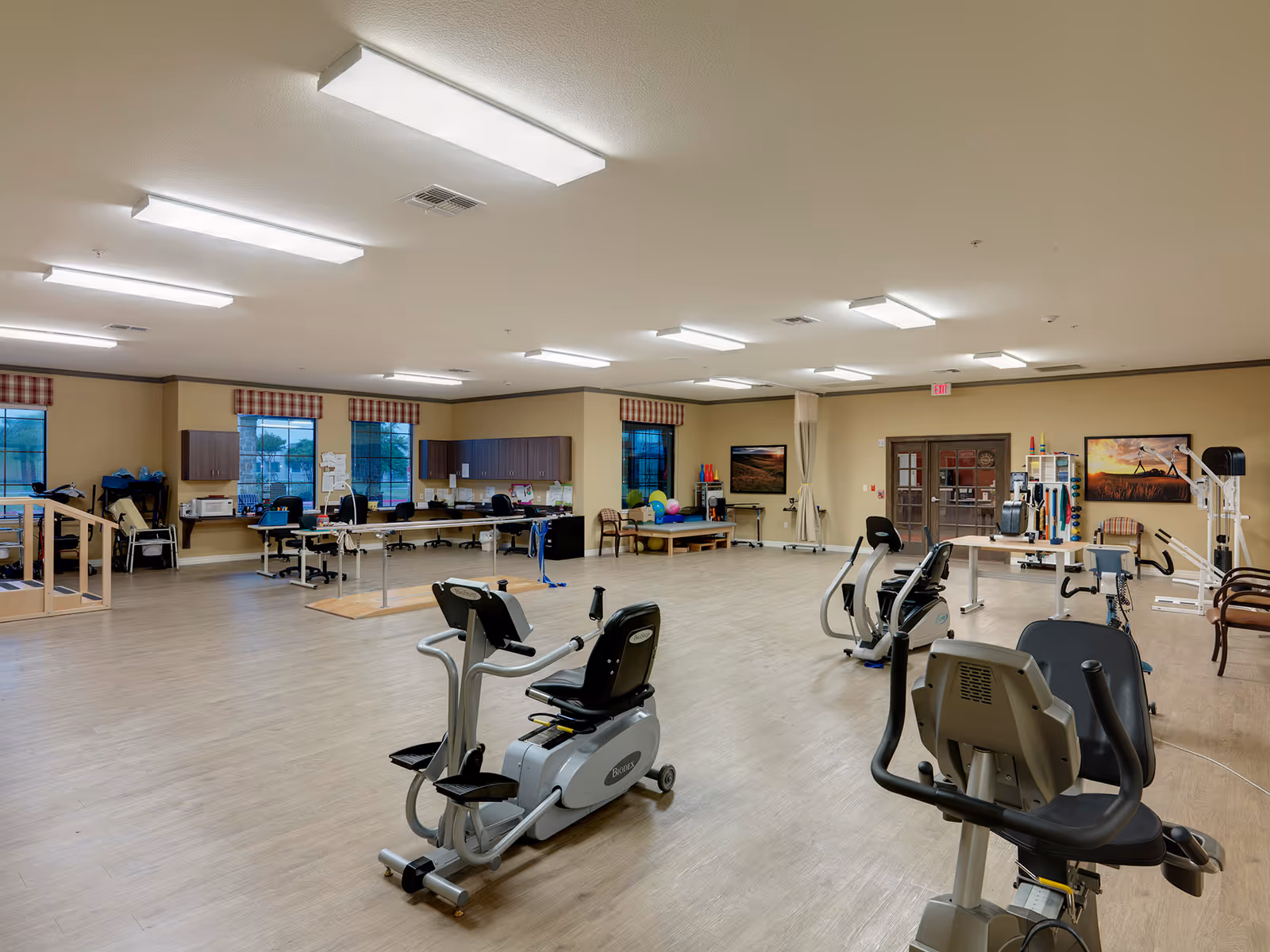 A spacious rehabilitation or physical therapy room with exercise equipment including stationary bikes and parallel bars. The room has wooden flooring, several desks and chairs along the walls, windows with plaid valances, and various therapy tools and equipment. The walls are painted beige and there are fluorescent lights on the ceiling.