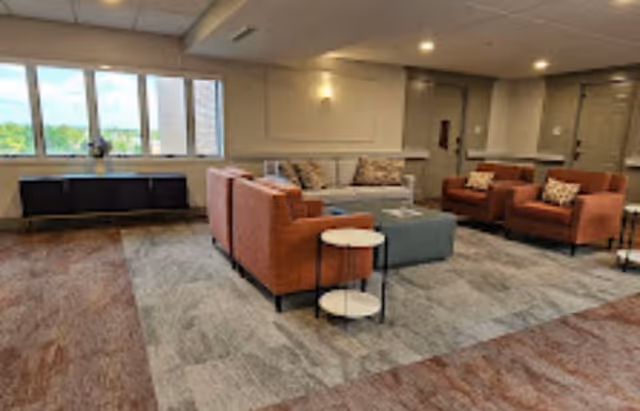 A spacious living room area with large windows letting in natural light. The room features a beige sofa, two orange armchairs, a blue ottoman, and a small round side table. The walls are light-colored with recessed lighting, and there is a dark wooden console table beneath the windows.