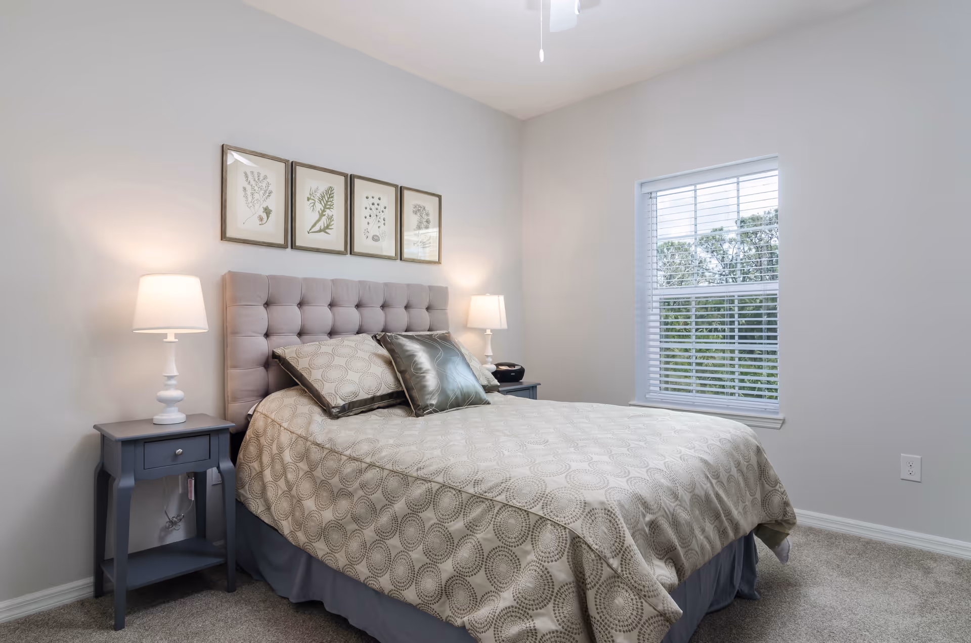 A neatly made bed with a tufted headboard in a bedroom. The bed has a beige patterned bedspread and two pillows, one beige and one metallic silver. There are two matching nightstands on either side of the bed, each with a white lamp. Above the headboard, four framed botanical prints are hung on the wall. A window with white blinds is on the right side of the room, letting in natural light and showing greenery outside. The walls are painted light gray and the floor is carpeted.