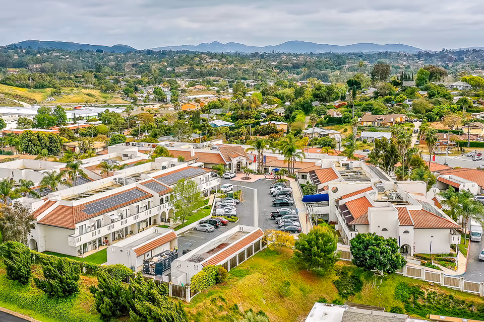 Aerial view of Rancho Vista Senior Living facility showing multiple white buildings with red-tiled roofs, parking lots with cars, surrounding greenery, and a suburban neighborhood in the background under a cloudy sky.