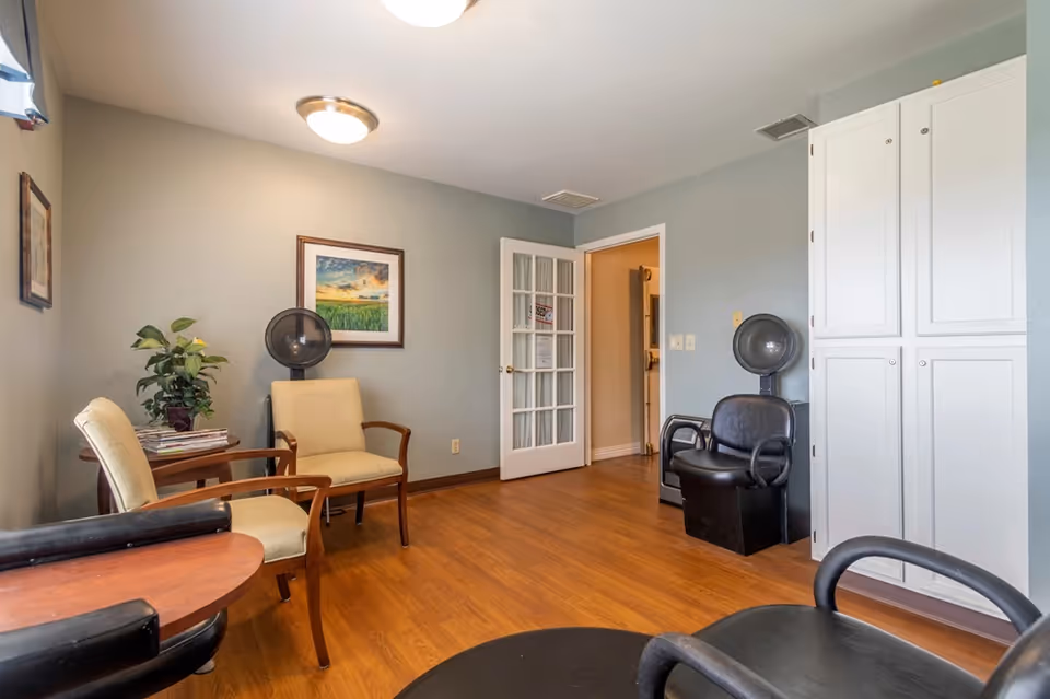 Small interior salon/common room with seating, two hooded hair dryers, a potted plant, framed art, and white storage cabinets.