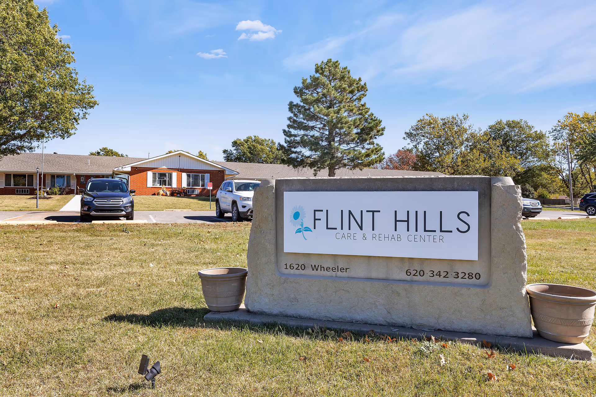 Stone entrance sign reading 'Flint Hills Care & Rehab Center' on a grassy lawn with the facility building and parked cars in the background.