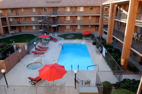 Outdoor swimming pool area in a senior living community with several red lounge chairs and umbrellas around the pool. There is a hot tub adjacent to the pool, surrounded by a fenced patio. The pool area is enclosed by a multi-story brick building with balconies overlooking the pool.