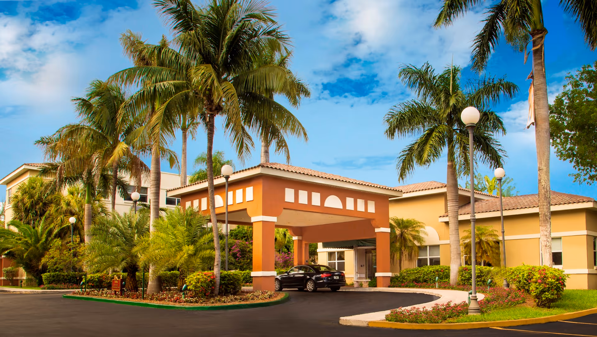 Exterior view of The Palace Nursing and Rehabilitation Center featuring a driveway with a covered entrance, surrounded by palm trees and well-maintained landscaping under a partly cloudy blue sky.