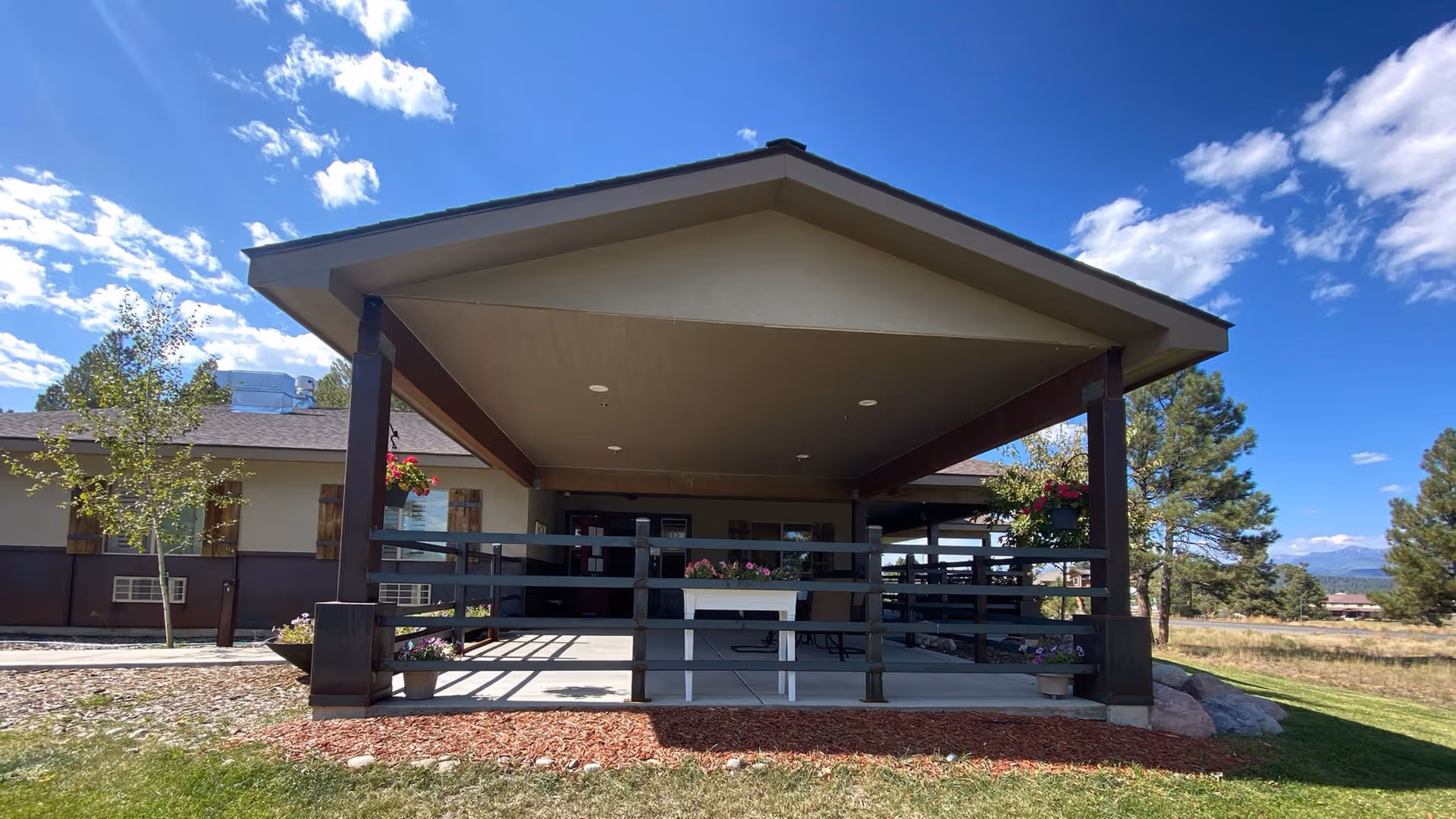 Covered entrance porch of a single-story senior living facility with a peaked roof, railings, and potted flowers under a blue sky.