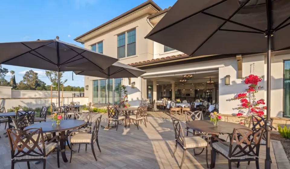 Outdoor patio area with multiple round tables and chairs under large umbrellas, adjacent to a building with large windows and an open dining area inside. The patio is decorated with flowers and overlooks a garden area with trees in the background under a clear blue sky.