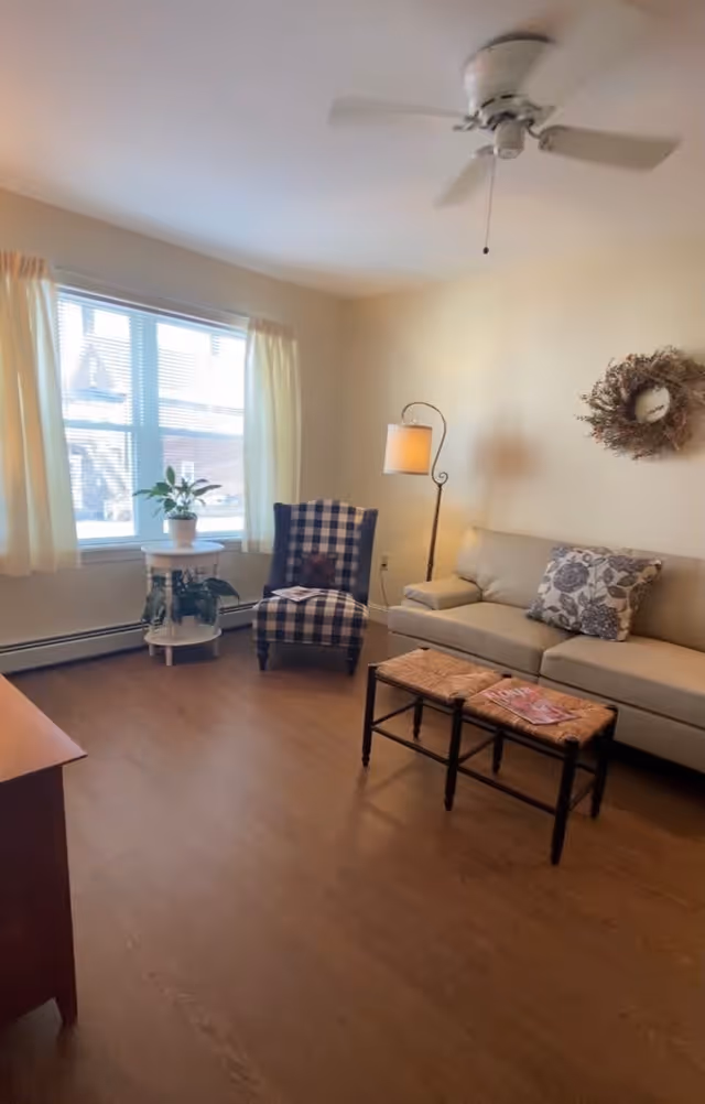 A cozy living room with a beige sofa adorned with a floral pillow, a checkered armchair, a small wooden coffee table with magazines, a floor lamp, a round side table with potted plants, a window with cream curtains, a ceiling fan, and a decorative wreath on the wall.