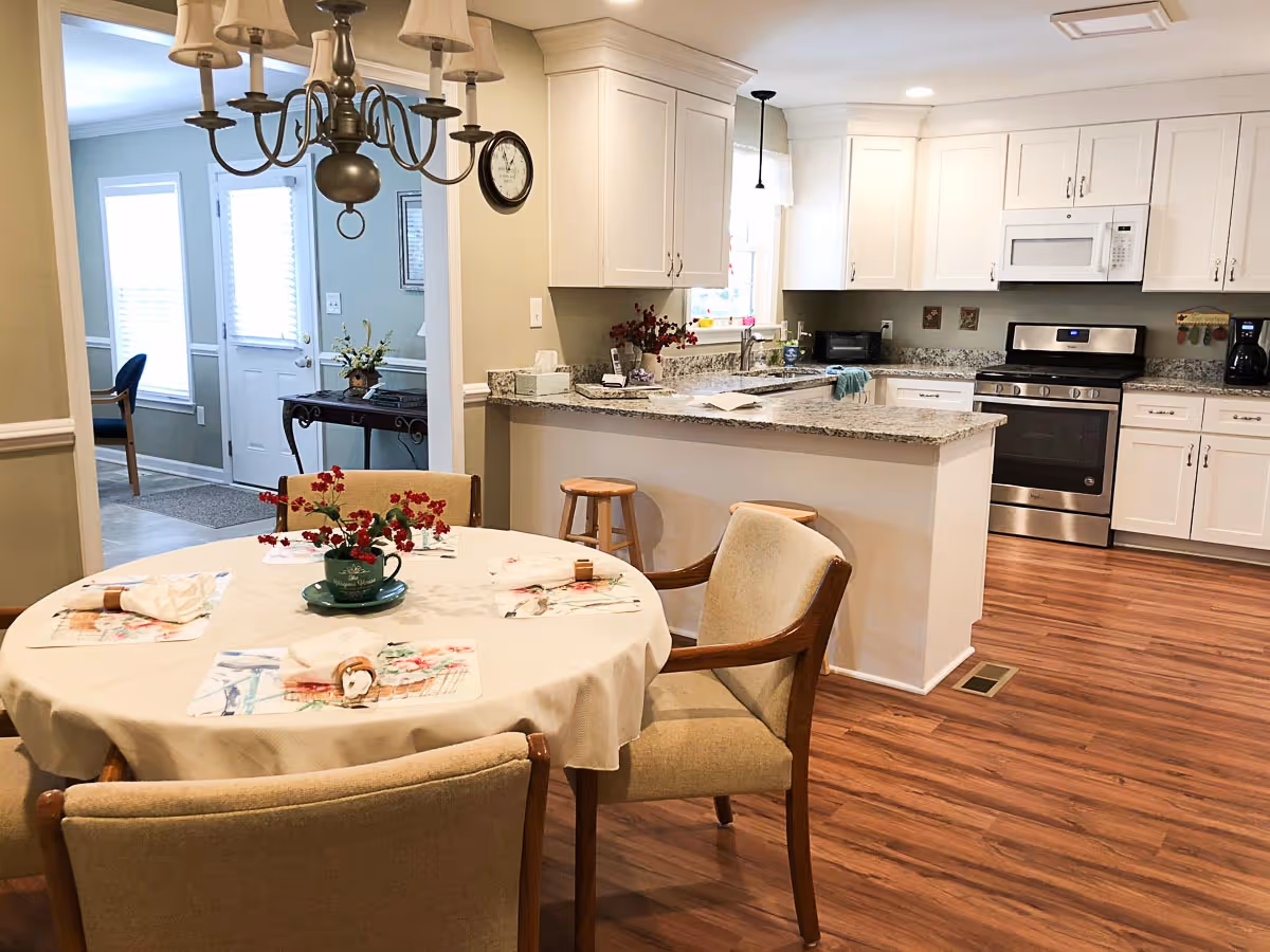 Bright open-plan dining area with a round table set for four and an adjacent white kitchen with granite countertops and stainless steel appliances.