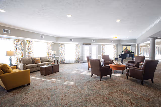 Sunlit communal lounge with sofas, armchairs, a central coffee table, and a grand piano near large windows.