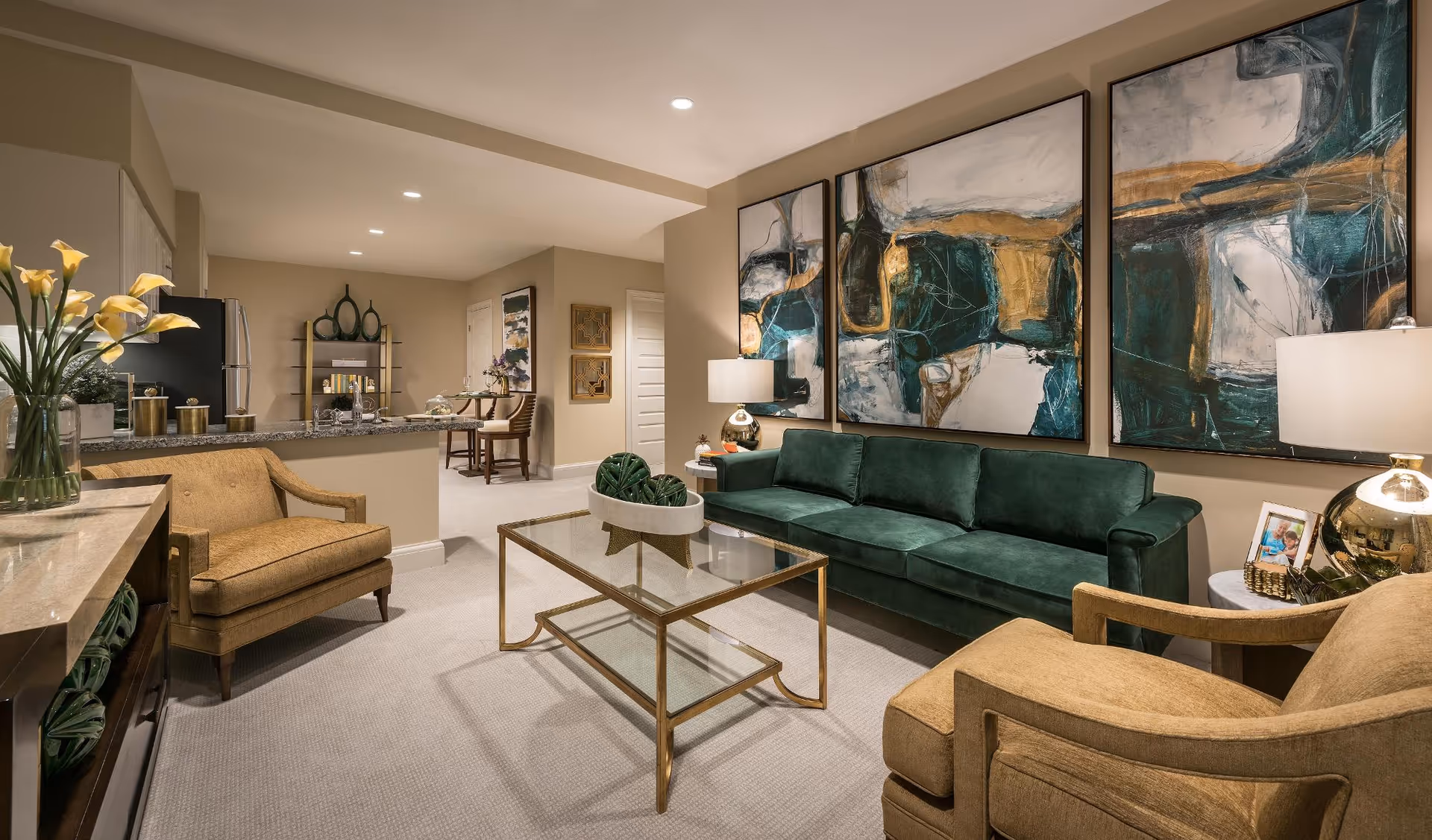 A modern living room with a green velvet sofa, two beige armchairs, a glass coffee table with a gold frame, and three large abstract paintings on the wall. The room has beige walls and carpet, with a kitchen and dining area visible in the background.