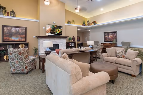 A spacious living room in an assisted living facility featuring beige sofas, a patterned armchair, a round brown ottoman, and a wooden coffee table. The room has a stone fireplace with a TV mounted above it, decorative plants, framed artwork on the walls, and soft overhead lighting.