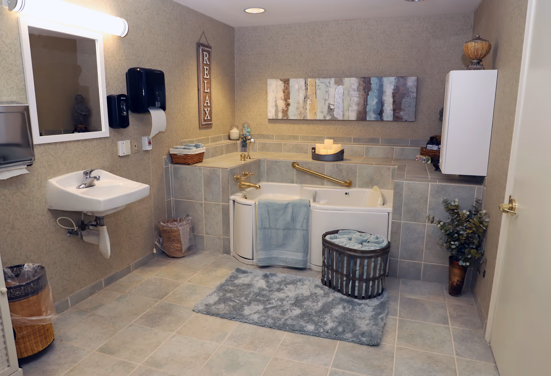 Accessible bathroom featuring a walk-in tub with grab bar, a wall-mounted sink and mirror, towels in a basket, and decorative accents.