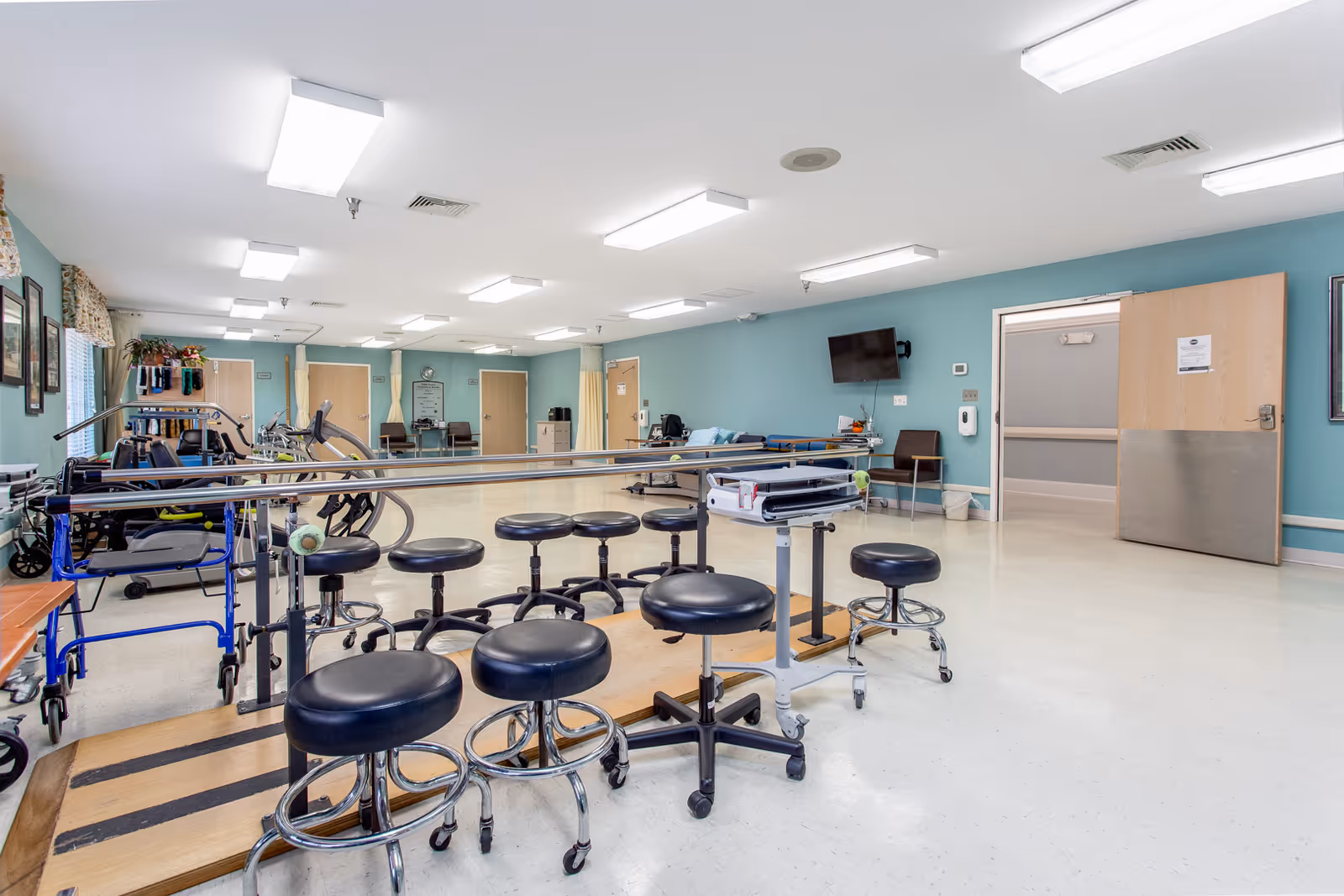 A spacious rehabilitation room with multiple black stools on wheels, parallel bars for walking exercises, exercise equipment, and chairs along the walls. The walls are painted light blue, and there are several doors and a wall-mounted TV visible.