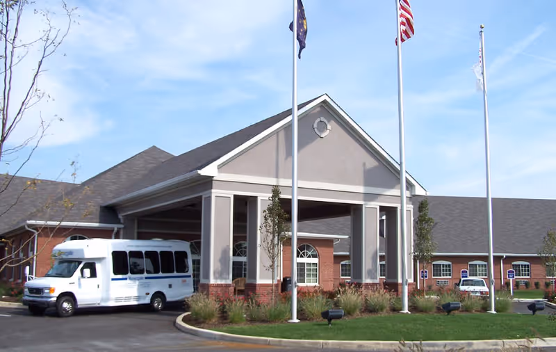 Exterior view of a senior living facility named Rosegate with a covered entrance supported by columns. There are three flagpoles with flags in front of the entrance, a white shuttle bus parked on the left side, and a well-maintained lawn with shrubs and small trees.