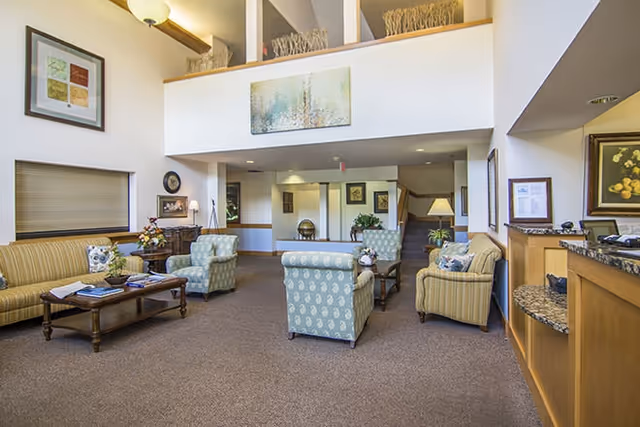 A spacious living room area in a senior living facility with multiple upholstered armchairs and sofas arranged around wooden coffee tables. The room features high ceilings with a balcony railing above, framed artwork on the walls, and a carpeted floor. There is a reception desk on the right side and decorative plants and lamps throughout the space.