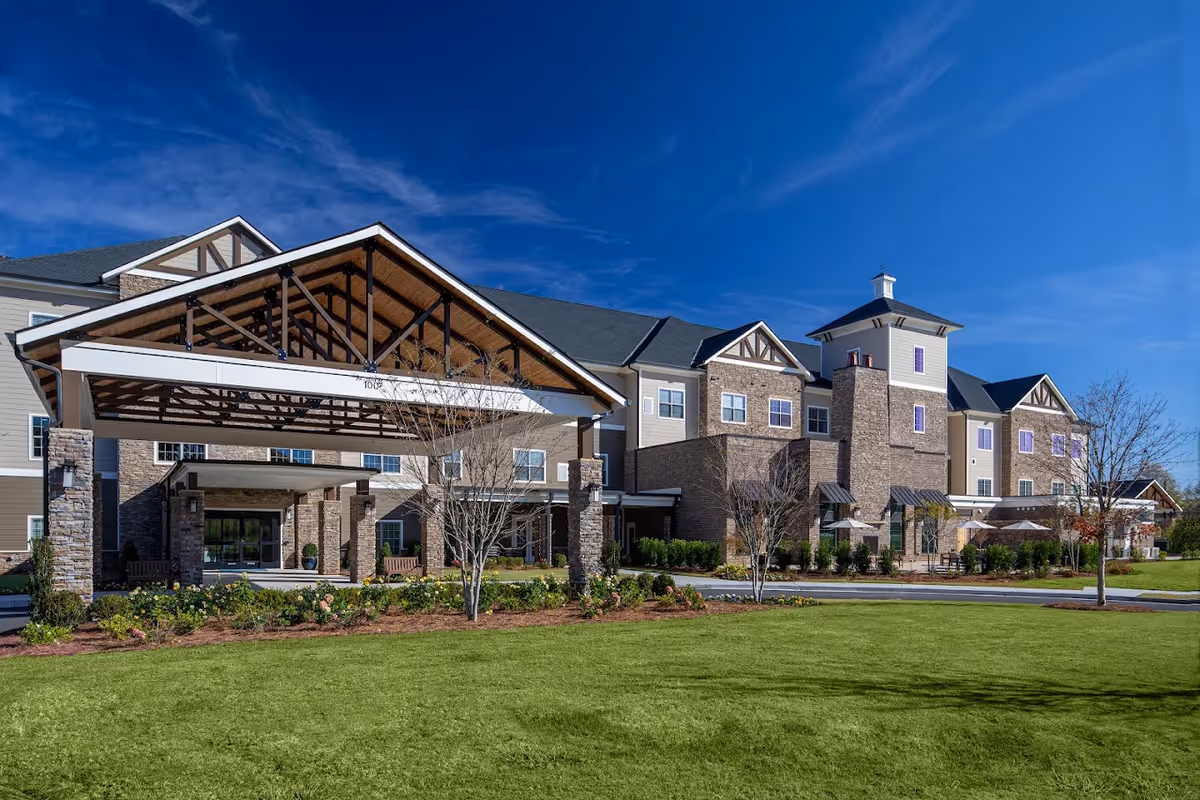 Exterior view of The Birches on Maple senior living facility showing a large building with stone and siding facade, a covered entrance with wooden beams, landscaped garden, and a clear blue sky.