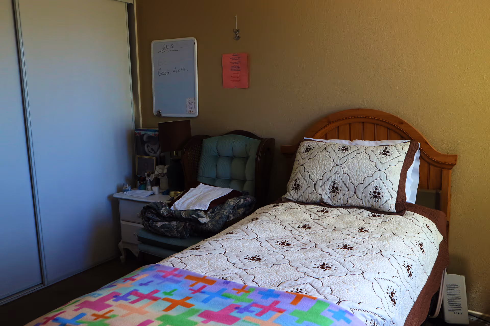 A small bedroom with a single wooden bed covered with a white quilt featuring a floral pattern and a colorful blanket at the foot. Next to the bed is a cushioned armchair with folded blankets on it, and a white nightstand holding a lamp and various small items. A whiteboard and a pink paper are attached to the beige wall above the chair. A sliding closet door is visible on the left side of the image.