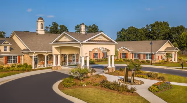 Exterior view of a senior living facility with a large covered entrance supported by white columns, surrounded by landscaped gardens and a circular driveway. The building has beige walls, red window shutters, and a brown roof with cupolas. Trees are visible in the background under a clear blue sky.