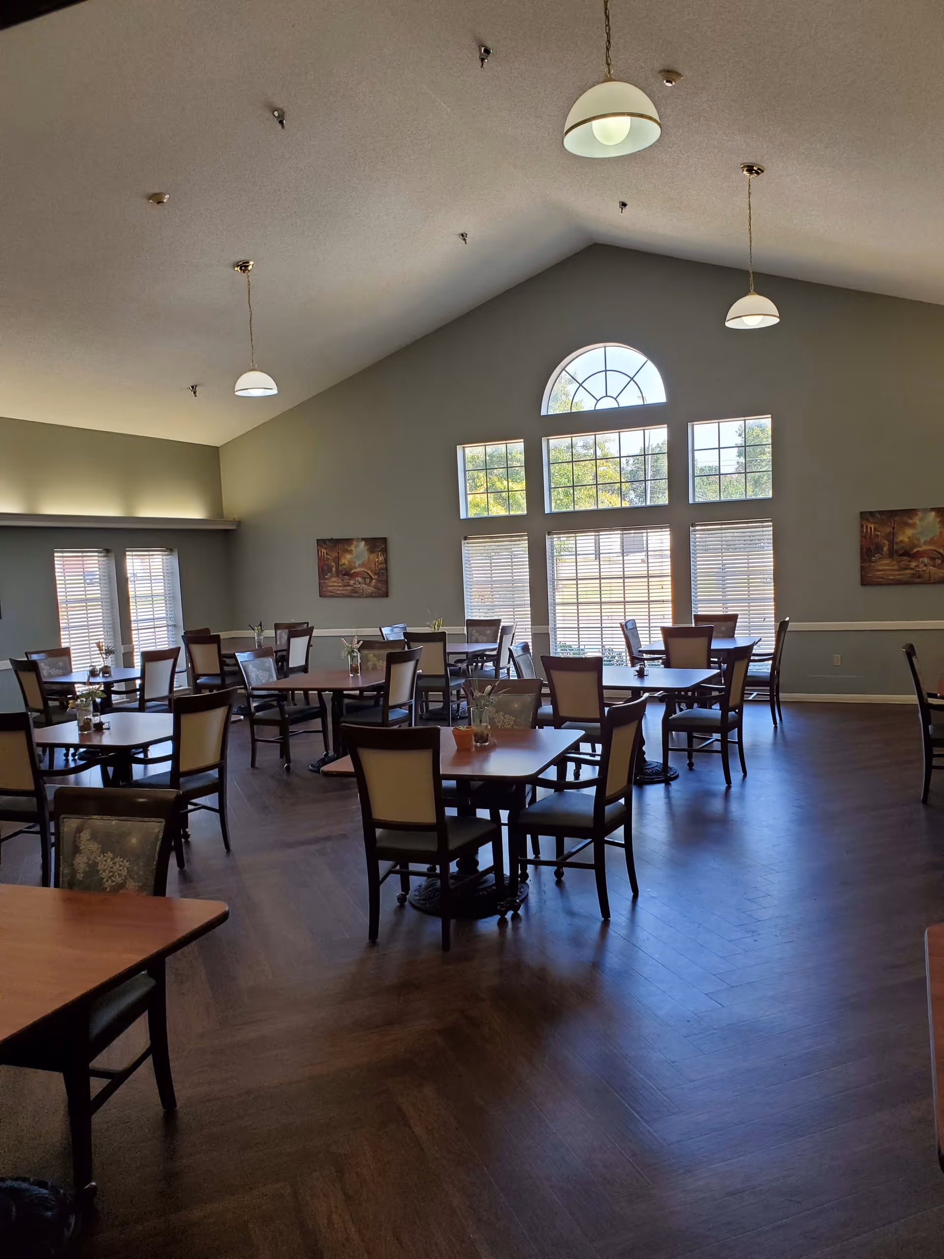 A spacious dining room with multiple wooden tables and chairs arranged neatly. Large windows with blinds allow natural light to fill the room. The ceiling is vaulted with hanging pendant lights, and there are paintings on the walls. The floor has a dark wood finish.