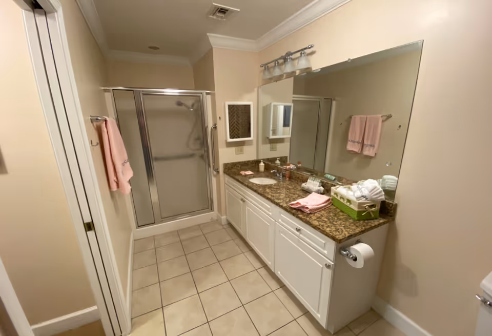Bathroom with tiled floor, a granite-top vanity and large mirror, pink towels, and a glass-enclosed shower stall.