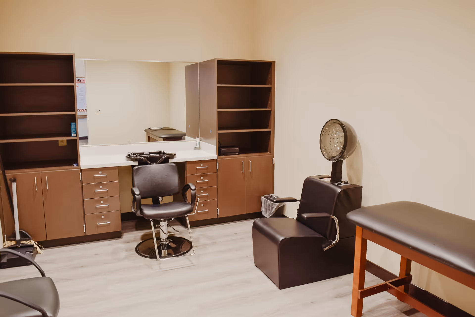 Interior view of a salon area in a senior living facility with a black salon chair in front of a large mirror, brown cabinets with shelves on either side, a black hair dryer chair, and a padded bench against a beige wall.