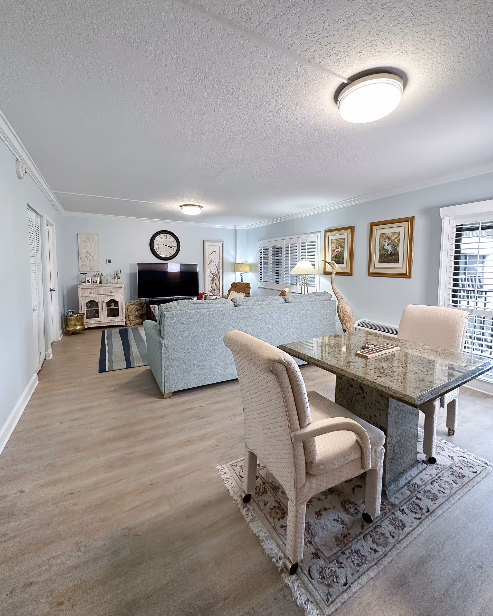 Open living area with pale blue sofas facing a TV, a marble-top dining table with upholstered chairs in the foreground, and framed artwork and lamps along the walls.
