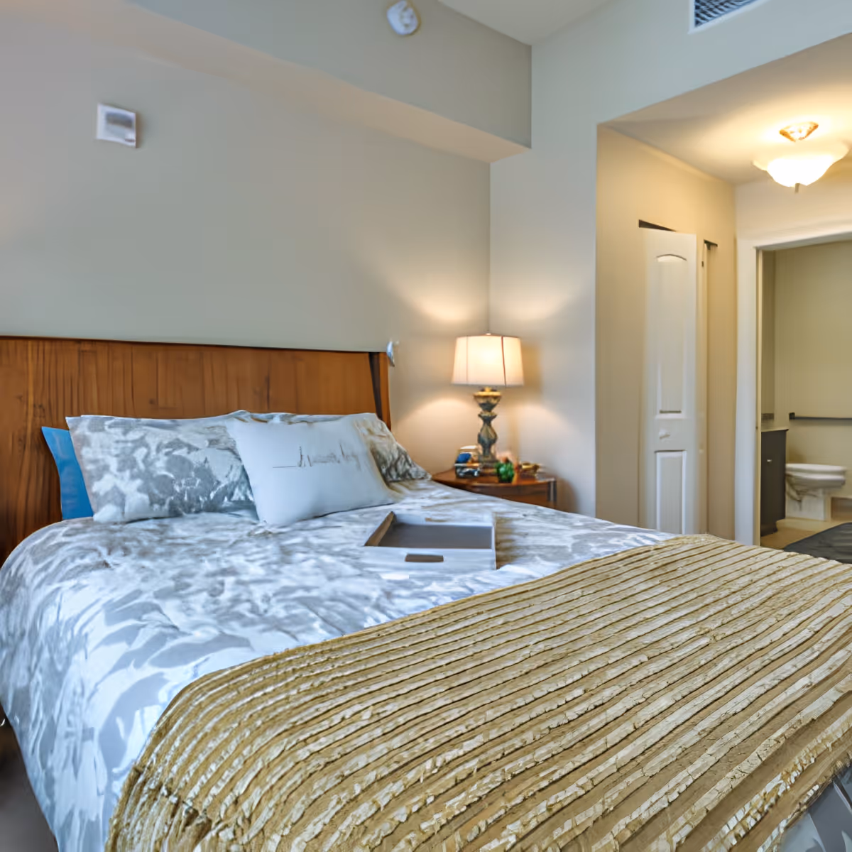 A bedroom with a wooden headboard, a bed with patterned bedding and pillows, a bedside table with a lamp, and an open door leading to a bathroom with a visible toilet.