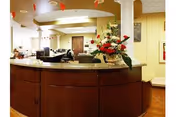 Reception area with a curved wooden desk topped with a granite counter, decorated with a large floral arrangement. The background shows a well-lit interior with beige walls, ceiling lights, and some seating areas.