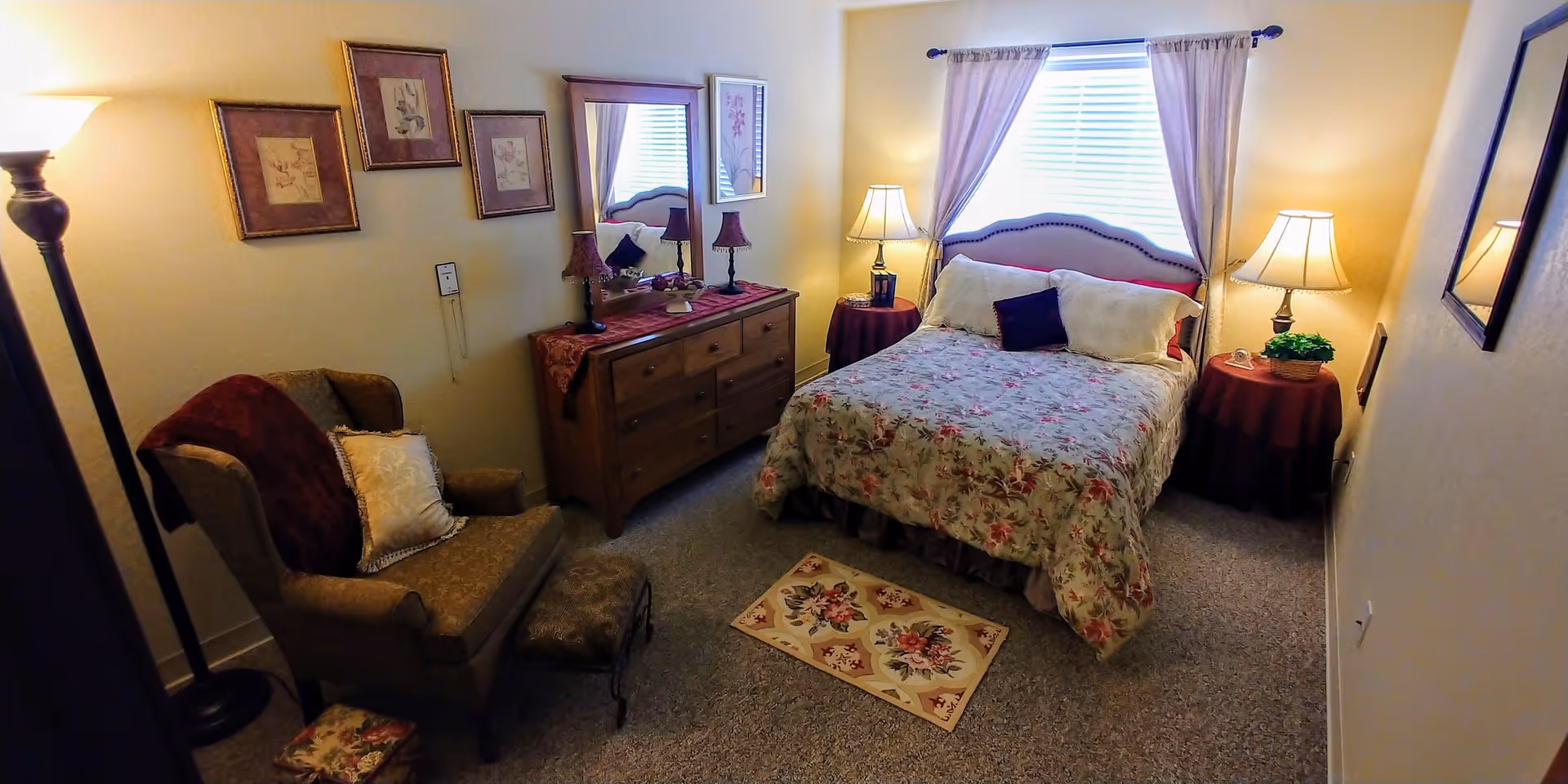 A cozy bedroom with a floral bedspread on a double bed, two bedside tables with lamps, a wooden dresser with a mirror and two small lamps, an upholstered armchair with a matching footstool, and framed artwork on the walls. The room is softly lit with a floor lamp and natural light coming through a window with curtains.
