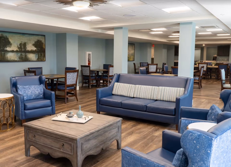A spacious common area in a senior living facility featuring blue leather sofas and armchairs arranged around a wooden coffee table. In the background, there are multiple dining tables with chairs, light blue walls, and framed landscape paintings. The flooring is wood, and the ceiling has recessed lighting.
