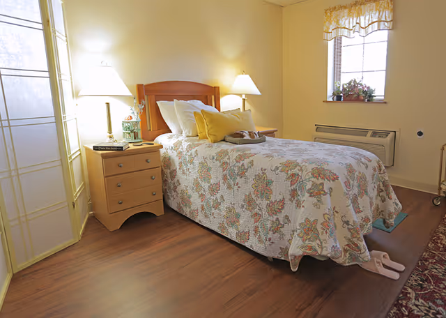 A cozy bedroom with a single bed covered in a floral quilt and multiple pillows. There are two bedside tables with lamps on each side of the bed. A small window with a floral valance lets in natural light. The room has wooden flooring and a pair of pink slippers placed near the foot of the bed.