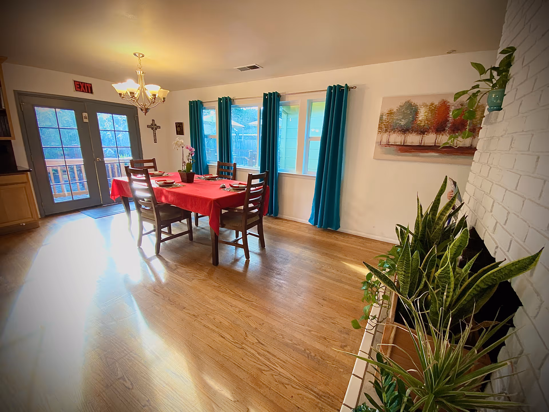 A bright dining room with a wooden floor, a table covered with a red tablecloth set for four people, teal curtains on the windows, a chandelier overhead, a white brick fireplace with several green plants, and a painting of trees on the wall.