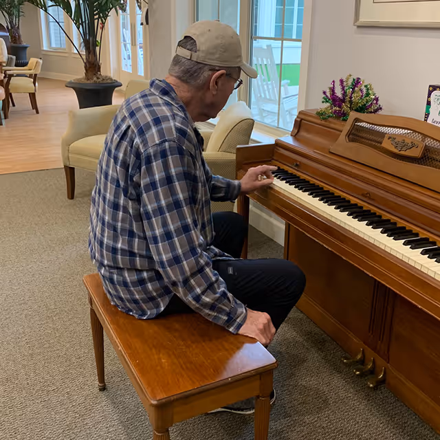 An elderly man wearing a plaid shirt and a beige cap is seated on a wooden bench playing an upright piano in a well-lit room with large windows and comfortable seating.