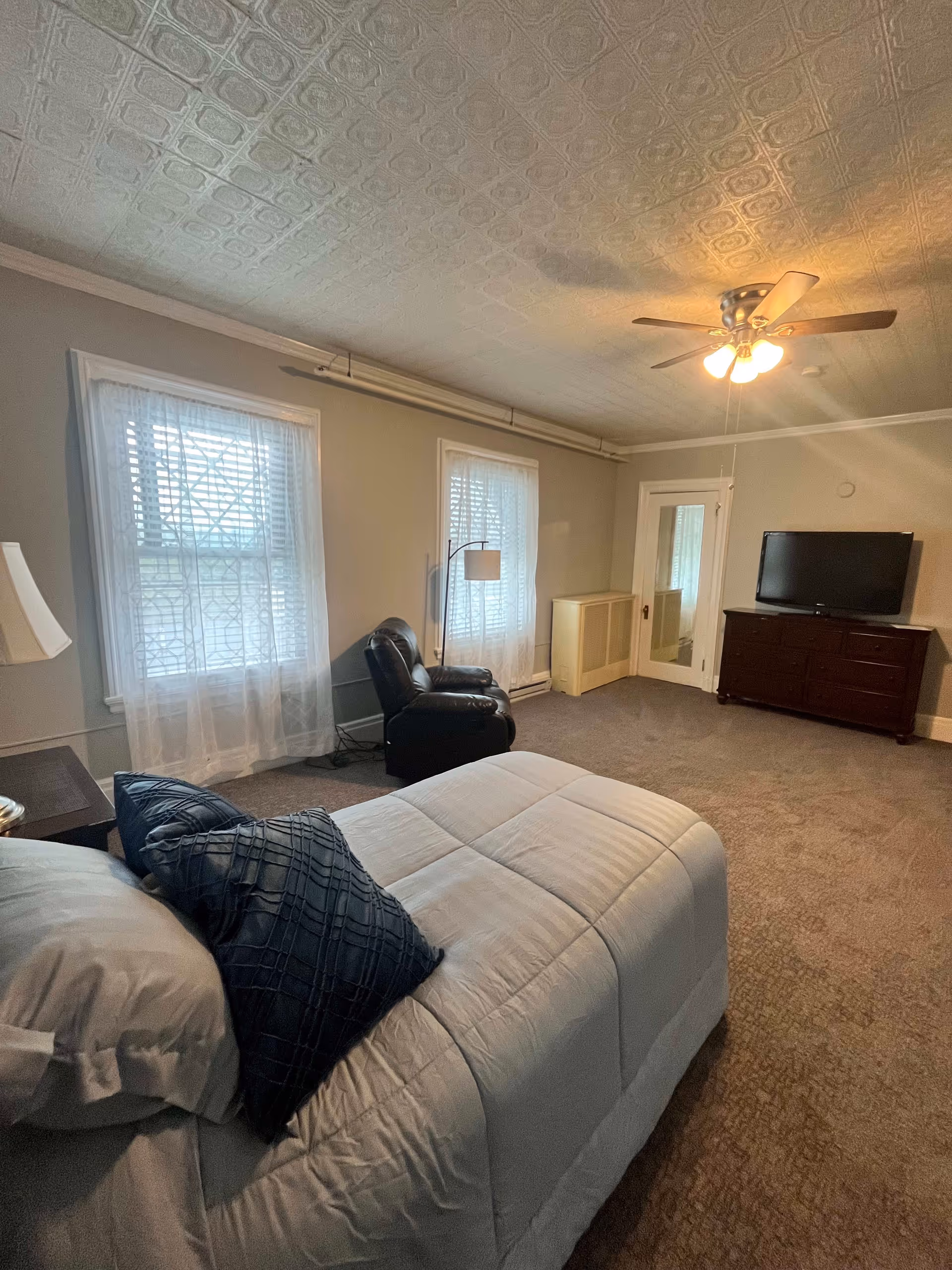 A bedroom in Westwind Manor featuring a bed with white bedding and dark blue pillows, two windows with sheer white curtains, a black recliner chair, a floor lamp, a ceiling fan with lights, a wooden dresser with a flat-screen TV on top, and a door with a mirror. The room has beige walls and carpeted flooring.