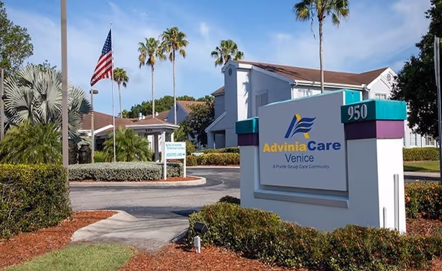 Exterior view of AdviniaCare Venice facility with a large sign displaying the facility name and address number 950. The scene includes palm trees, an American flag on a flagpole, well-maintained landscaping, and a clear blue sky.