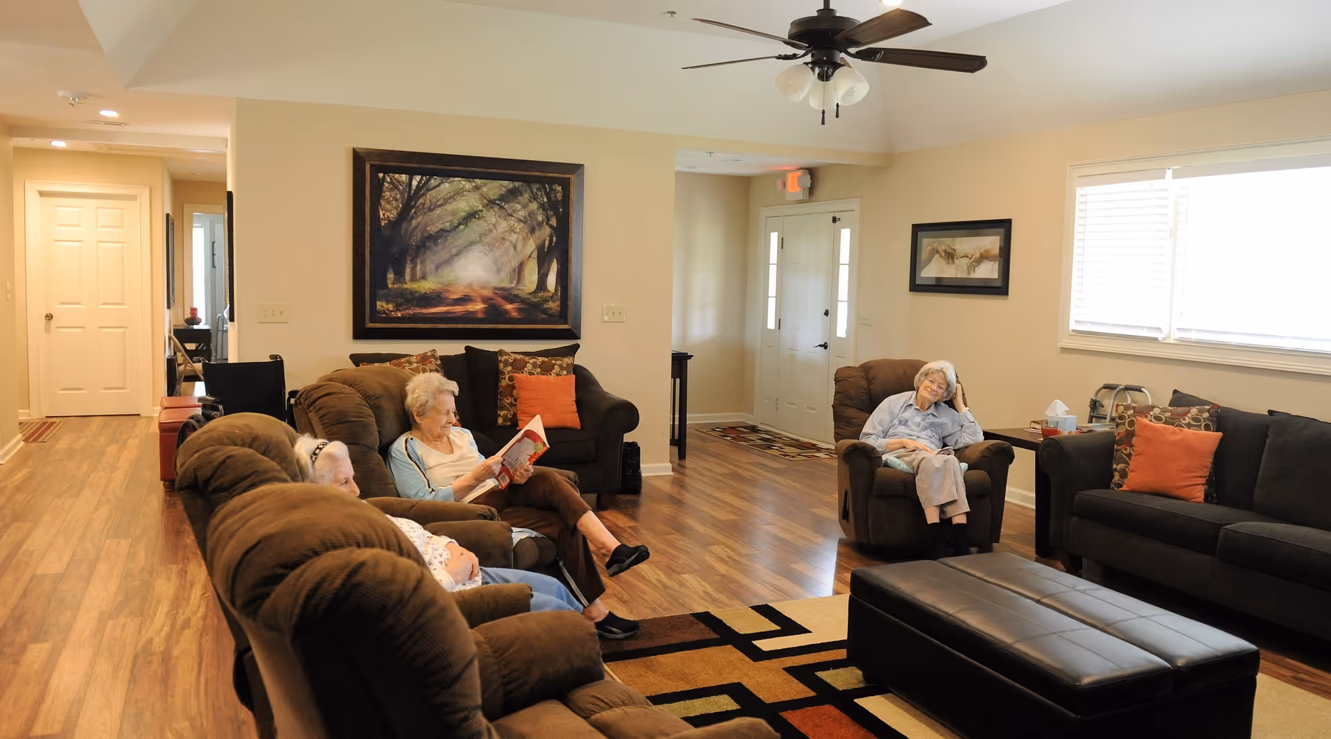 A cozy living room in a senior care facility with three elderly women seated on brown cushioned chairs and sofas. One woman is reading a book, while another is resting with her eyes closed. The room has wooden flooring, a ceiling fan, framed artwork on the walls, and a large window with blinds letting in natural light. There is a patterned area rug and a black leather ottoman in the center.