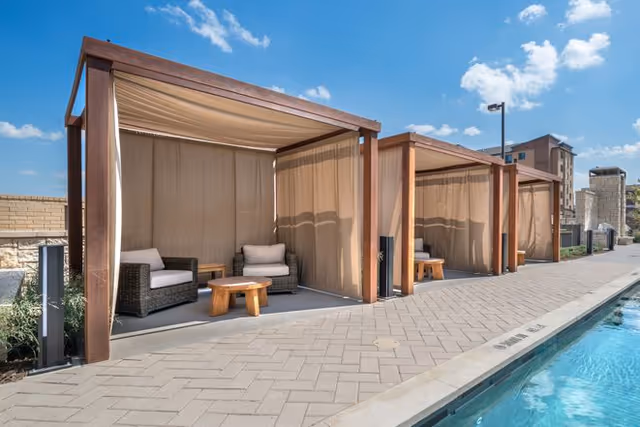 Outdoor poolside cabanas with wicker chairs and small wooden tables under wooden pergolas with beige curtains, next to a swimming pool on a sunny day with a blue sky and scattered clouds.