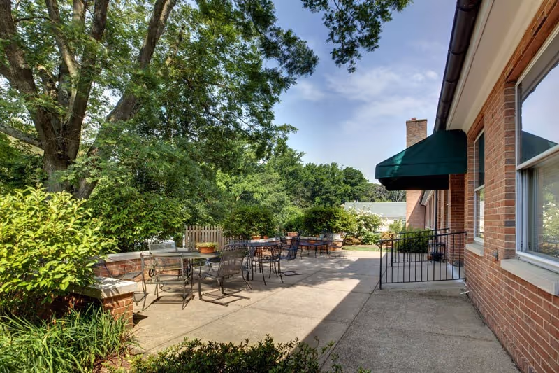 Sunny outdoor patio with metal tables and chairs next to a brick building and large trees.