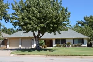 Single-story residential building with a large tree in the front yard, a well-maintained lawn, and a driveway leading to a two-car garage under a clear blue sky.