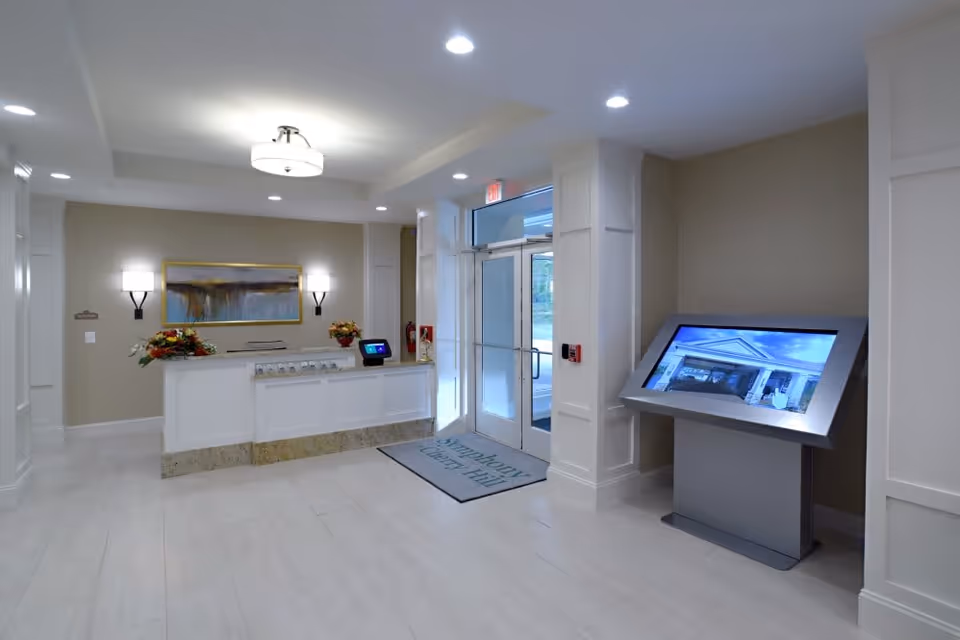 Reception area of a senior living facility with a white front desk adorned with flower arrangements, a large painting on the wall, and modern lighting fixtures. There is a glass door entrance with a mat that reads 'Symphony Cherry Hill' and a large touchscreen display on a stand to the right.