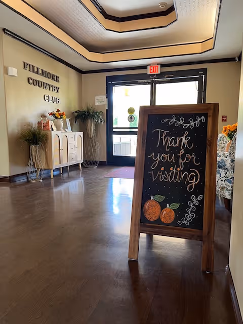 Entrance lobby of Fillmore Country Club showing a chalkboard sign reading 'Thank you for visiting,' decorative furniture, and the front doors.