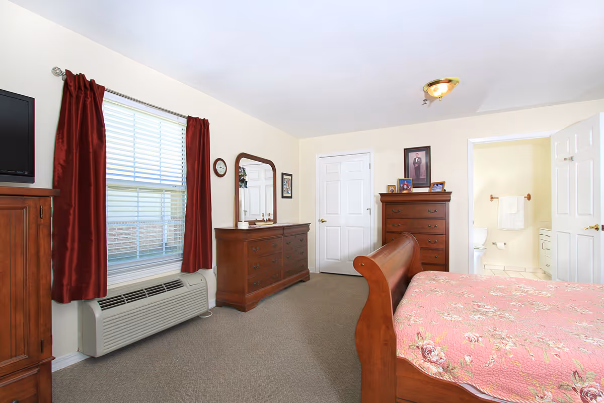 A bedroom with a wooden bed frame covered with a pink floral quilt. The room has beige walls and carpeted floor. There is a wooden dresser with a mirror, a tall chest of drawers with framed photos on top, and a window with red curtains. A door leads to a bathroom with a visible toilet and towel rack.